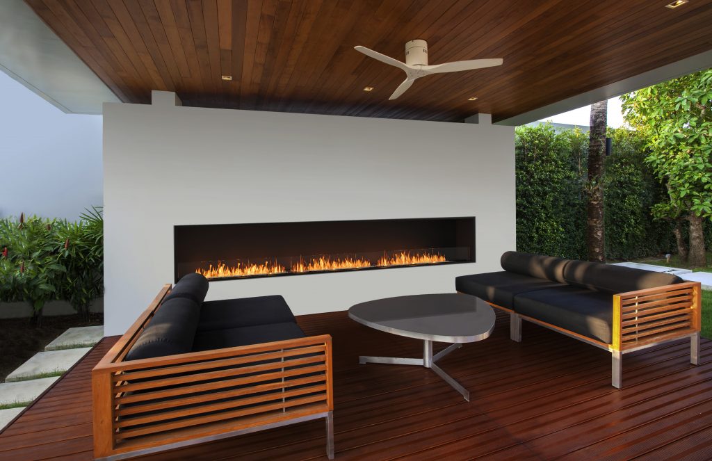 Interior and exterior design of outdoor cover space with wooden floor and wooden ceiling which features ceiling fan, lighting, middle round table, sofa and black cushion