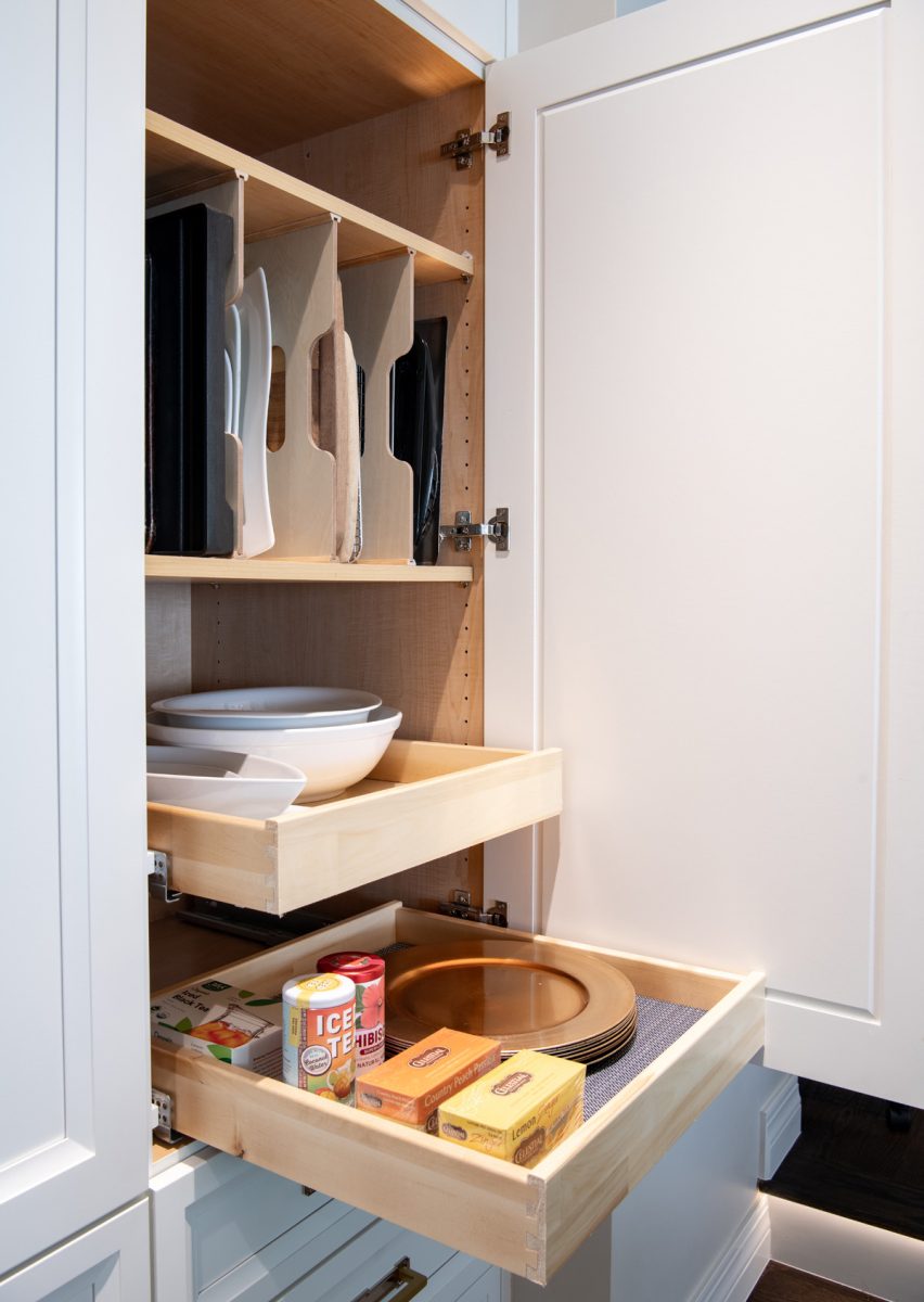 Open kitchen cabinet with pull-out shelves holding cutting boards, bowls, plates, and tea boxes.